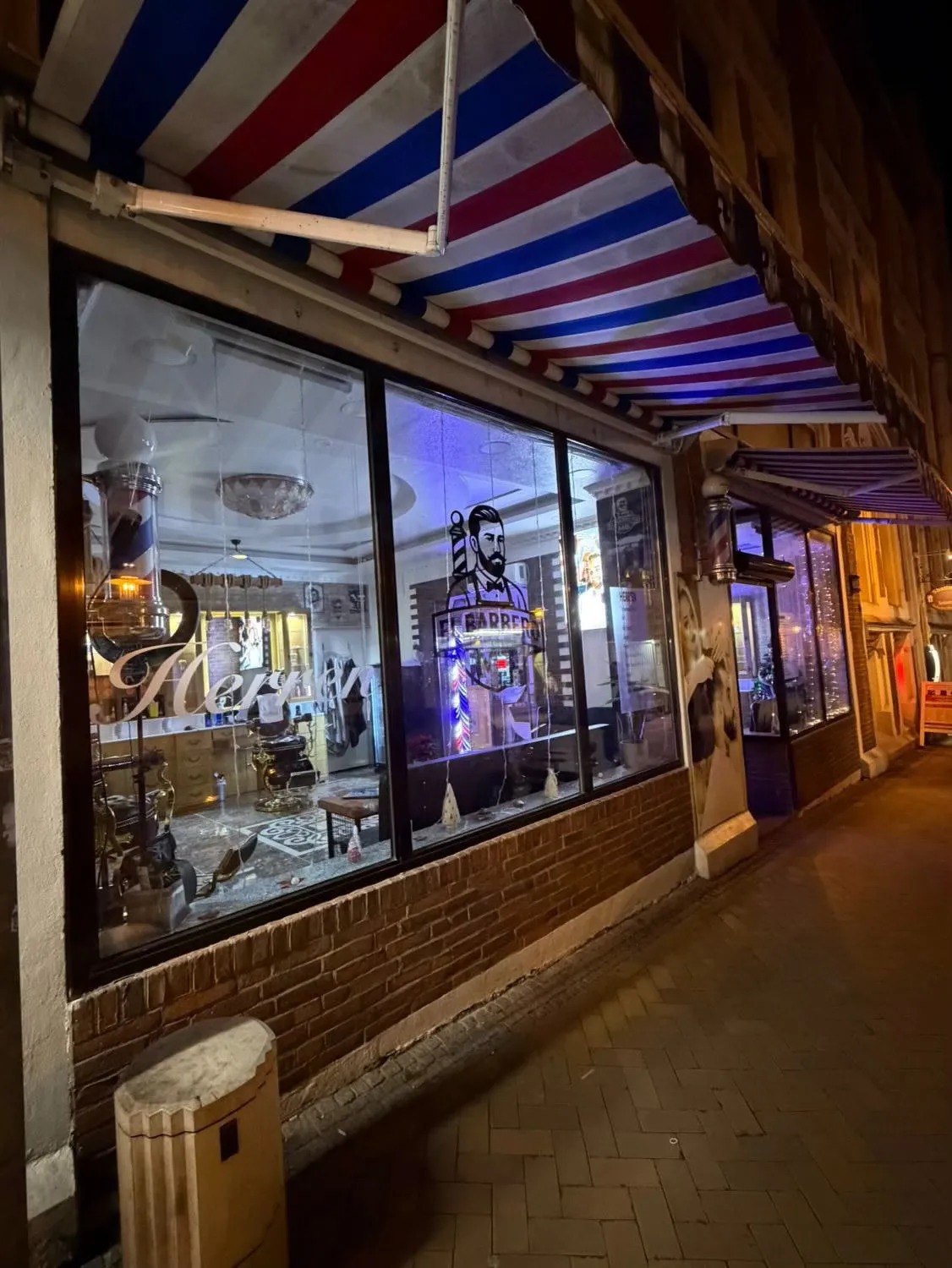 El barbero MB - Haar- und Hautpflegebehandlungen - Nachtansicht einer Friseursalonfassade mit einem beleuchteten Neonschild mit einem bärtigen Mann, einer gestreiften Markise und einem durch das Glas sichtbaren, gemütlichen Innenraum.